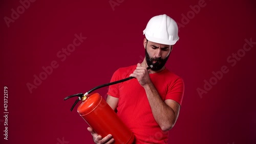 the man doesn't know what to do with the fire extinguisher and throws it away, he wears a safety helmet, wears a red T-shirt, bearded, dark-haired, background red