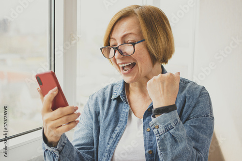 Wow. Excited senior mature woman surprised by good unbelievable news, holding and looking at mobile phone delighted clench fist victory triumph gesture, feeling amazed. Unexpected win, sales offer 