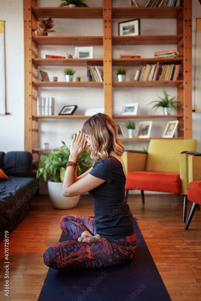 Side view of a fit woman sitting on a yoga mat at home in the lotus position and meditating. Side view of a fit woman sitting on a yoga mat at home in the lotus position and meditating.