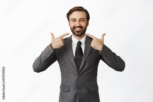 Handsome successful businessman pointing at his white smile, showing perfect healthy teeth, standing in suit over white background