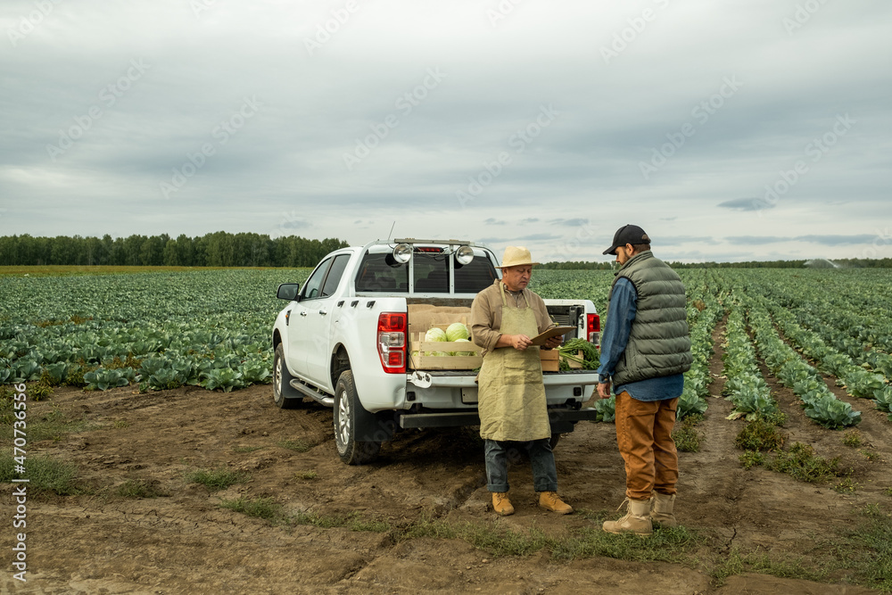 Mature Asian vegetable farmer in sunhat and apron reading papers into clipboard while talking to customer on plantation - obrazy, fototapety, plakaty 