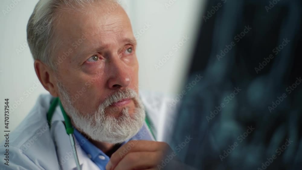 Close-up face of focused mature male doctor examining head MRI scan of patient sitting at desk in medical office room, thinking about diagnosis, expressing concern of illness. Shooting in slow motion. Close-up face of focused mature male doctor examining head MRI scan of patient sitting at desk in medical office room, thinking about diagnosis, expressing concern of illness. Shooting in slow motion.