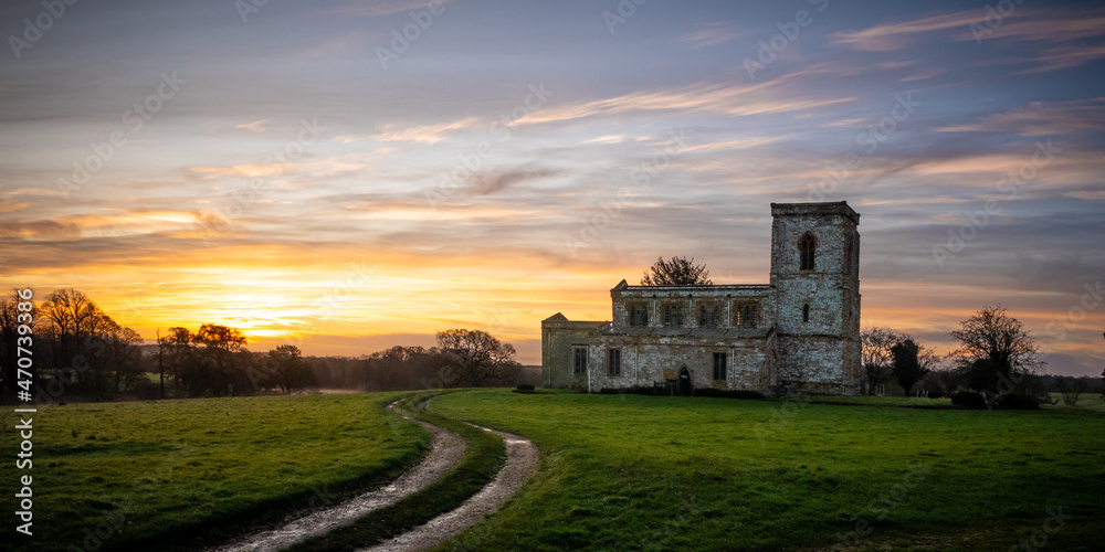 An ancient English church stands in a meadow against a backdrop of a magnificent sunrise. In the foreground a set of tractor tyre tracks leads the eye through the field.