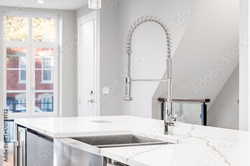 A kitchen sink detail shot with a chrome faucet, stainless steel apron sink, and white granite counter tops looking out towards a staircase and empty living area Fototapeta