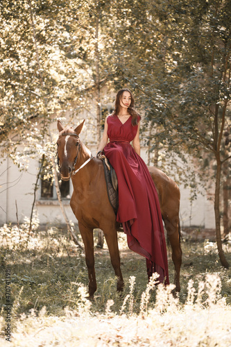 A beautiful young woman in a silk dress sits astride a horse. Horse ride on a sunny day in the forest.