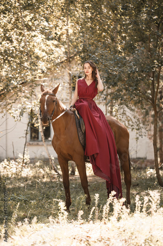 A beautiful young woman in a silk dress sits astride a horse. Horse ride on a sunny day in the forest.