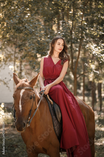 A beautiful young woman in a silk dress sits astride a horse. Horse ride on a sunny day in the forest. © Дмитрий Ткачук