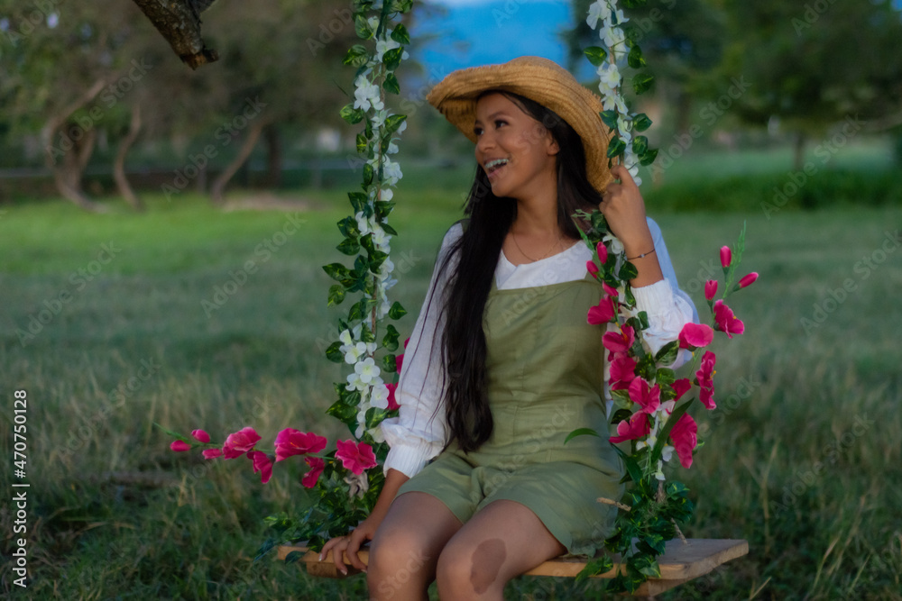 Beautiful smiling teenage woman with long black hair, caucasian brunette, with hat sitting on a swing with flowers in the forest.