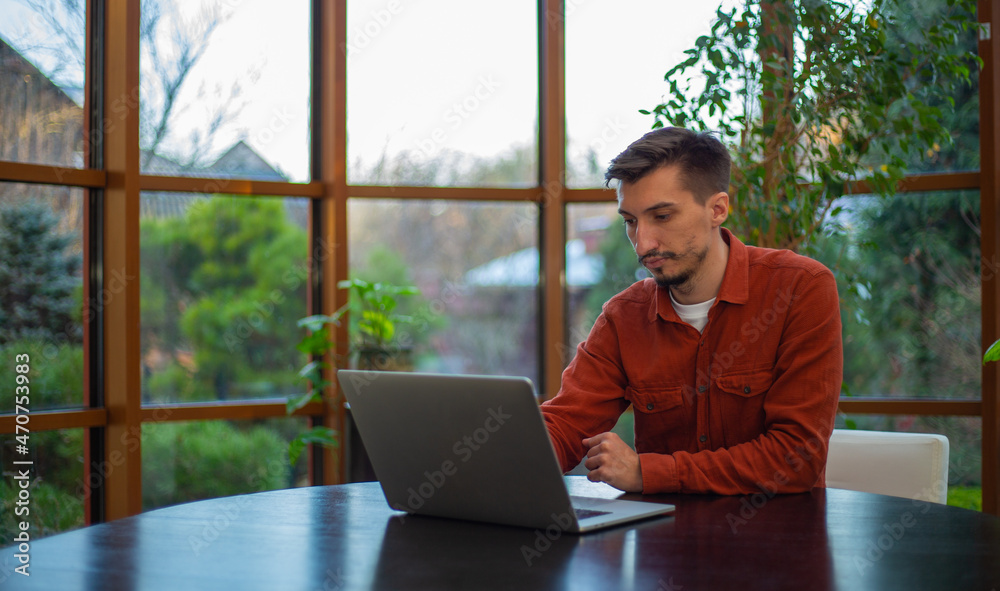 A man in a red shirt works at a laptop at a table during the day against the background of the garden. A man in a red shirt works at a laptop at a table during the day against the background of the garden.