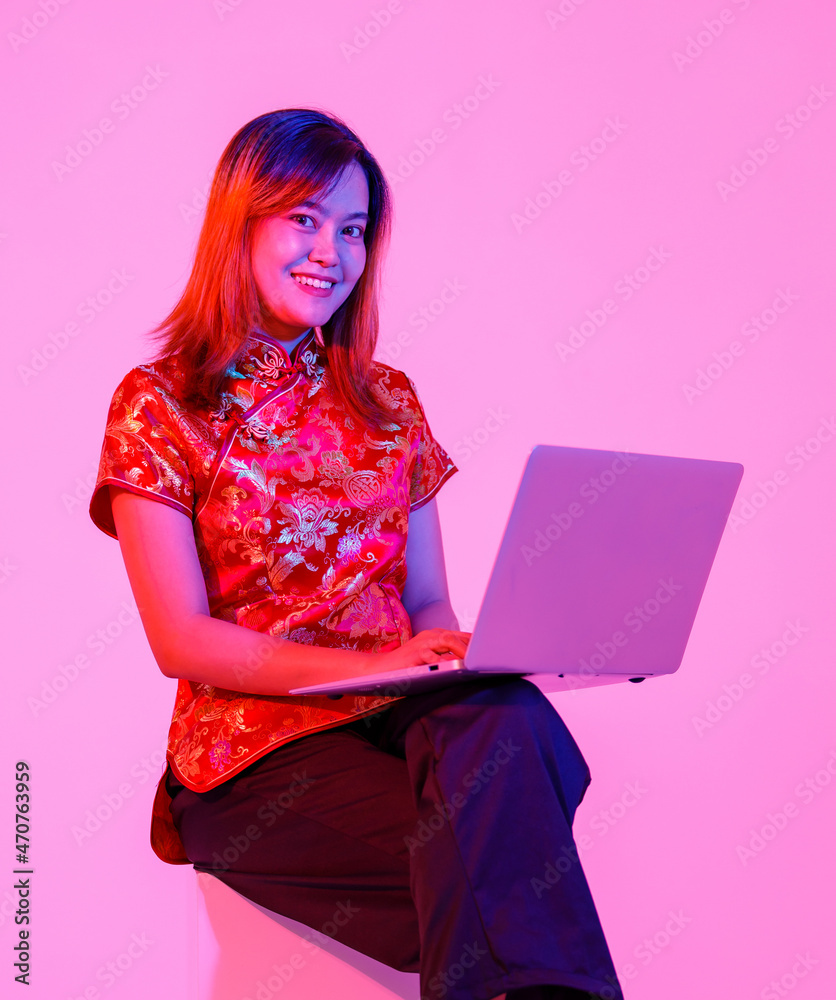Studio shot of millennial Asian young teen female model in beautiful red Chinese traditional cheongsam qipao shirt sitting holding laptop computer surfing shopping online on pink light background Studio shot of millennial Asian young teen female model in beautiful red Chinese traditional cheongsam qipao shirt sitting holding laptop computer surfing shopping online on pink light background