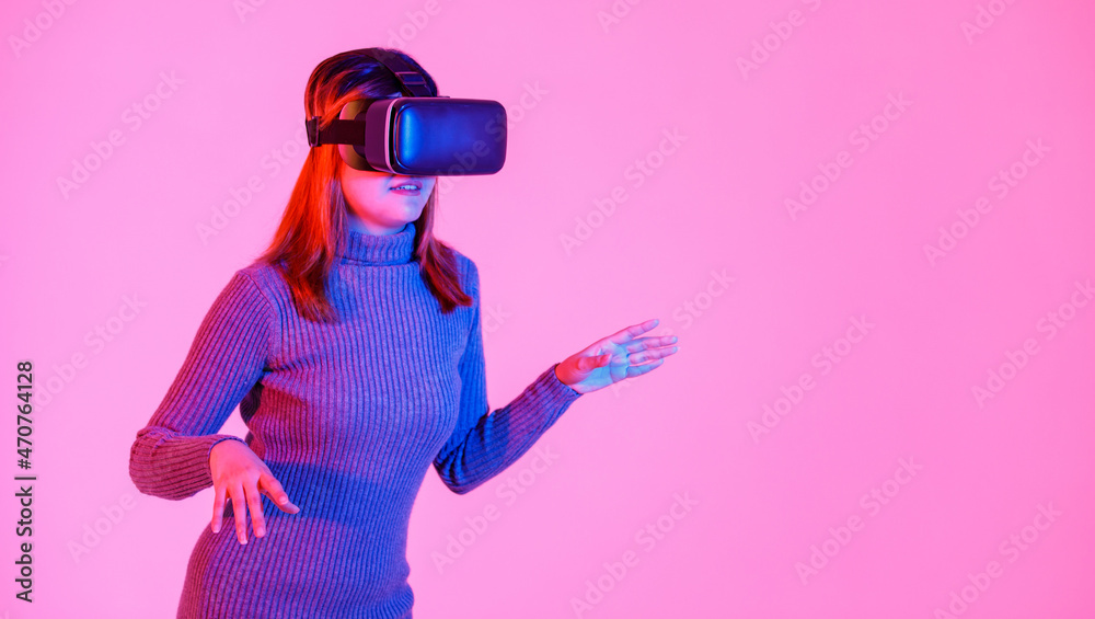 Studio shot of Asian young teen female model in gray turtleneck dress wears artificial intelligence virtual reality vr video game goggles headset playing 3d console gameplay on pink light background