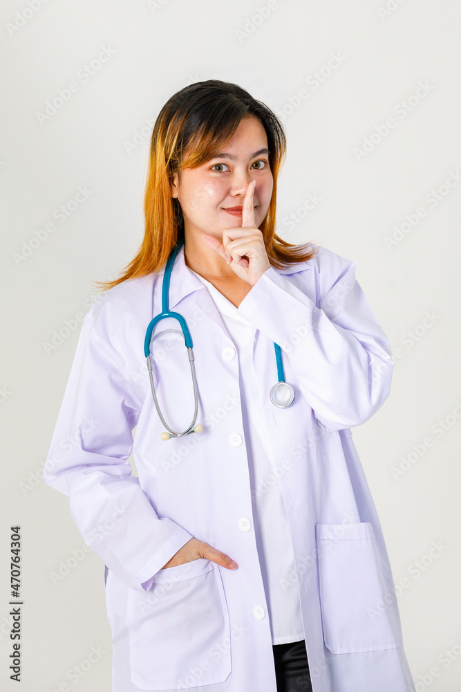 Portrait studio shot of Asian professional female hospital doctor in lab coat uniform with stethoscope standing smiling holding index finger at mouth tell to quiet mute silent on white background Portrait studio shot of Asian professional female hospital doctor in lab coat uniform with stethoscope standing smiling holding index finger at mouth tell to quiet mute silent on white background