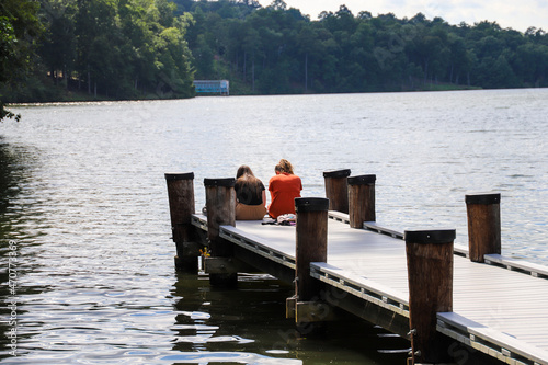 two girls sitting on the edge of a long brown wooden dock over the still blue lake waters surrounded by lush green trees and clouds at Lake Kedron in Peachtree City, Georgia USA photo