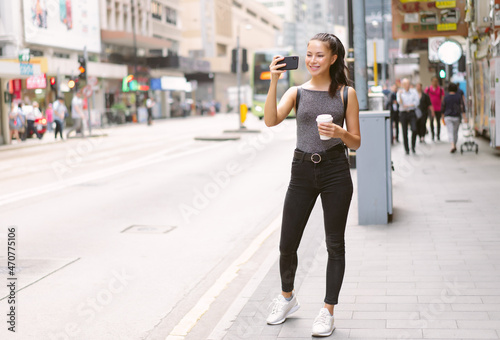 Happy beautiful woman holding coffee hot cup and taking photo with mobile phone in Central walking in streets, travel guide, tourism in Hong Kong. Young girl tourist with smartphone on the street.