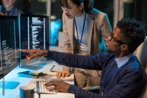 Two young intercultural colleagues discussing coded data on computer screen while African businessman pointing at it