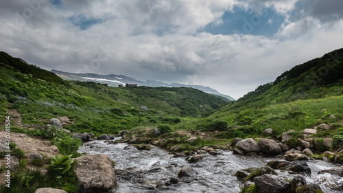 The stream flows through a rocky bed in the valley. Green grass and wildflowers on the banks and mountain slopes. Clouds in the blue sky. Kamchatka