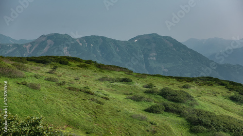 Green vegetation is visible on the hillside: grass and shrubs. A picturesque mountain range against the sky. Kamchatka.