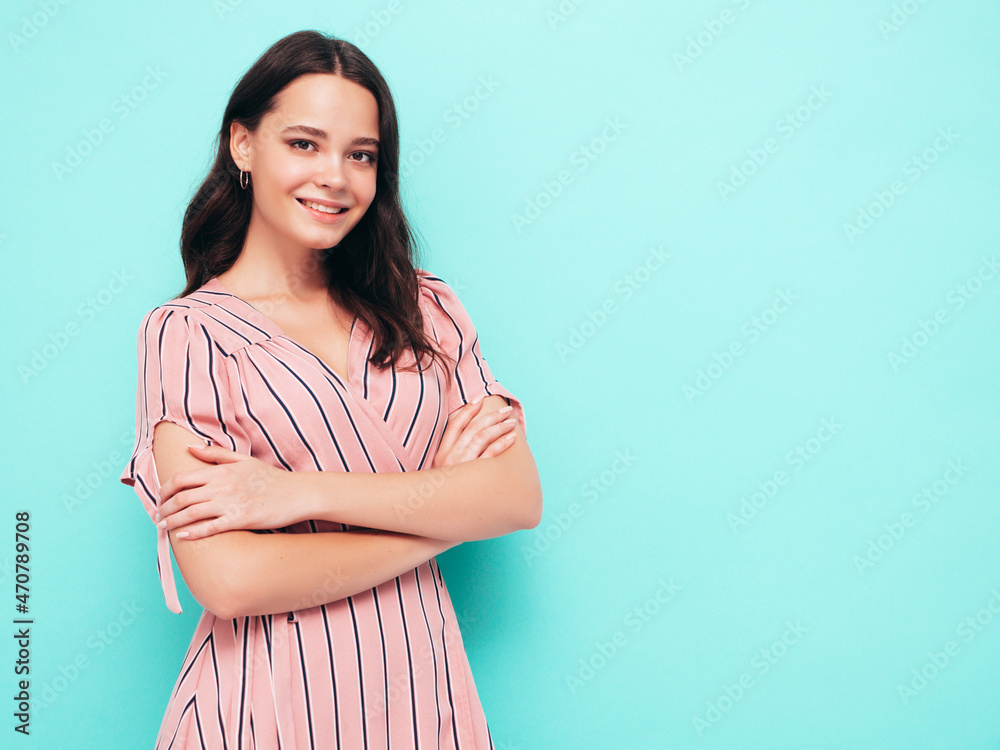Obraz premium Portrait of young beautiful smiling female in trendy summer pink dress. Sexy carefree woman posing near blue wall in studio. Positive model having fun indoors. Cheerful and happy