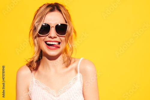 Young beautiful smiling female in trendy summer clothes. Sexy carefree woman posing near blue yellow in studio. Positive model having fun. Cheerful and happy. In sunglasses. Shows tongue