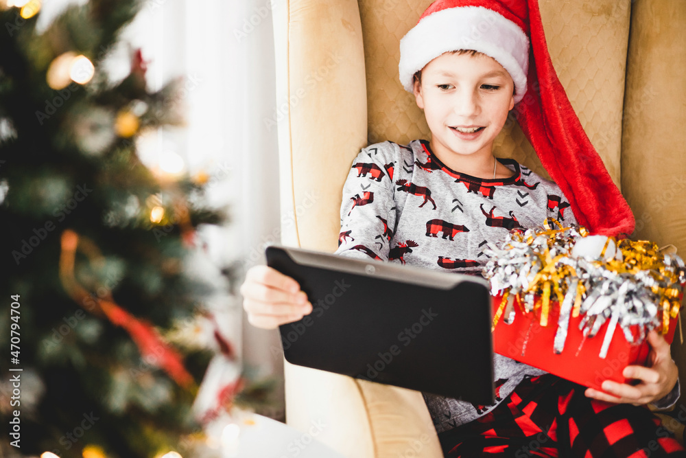 Happy teenager boy in pajamas and red santa hat with tablet having video chat at home. Smiling child sitting on armchair watching video at laptop. Christmas tree with decorative toys and lights. Happy teenager boy in pajamas and red santa hat with tablet having video chat at home. Smiling child sitting on armchair watching video at laptop. Christmas tree with decorative toys and lights.
