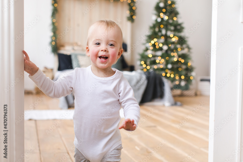 Adorable infant standing in doors looking at camera on background of Christmas tree in bedroom. Adorable infant standing in doors looking at camera on background of Christmas tree in bedroom.