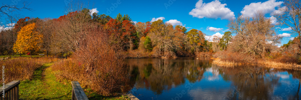Peaceful autumn forest landscape over the serene pond with green footpath. Blue sky, white clouds, and reflections of colorful tree foliage on the calm water of the lake in New England, USA.