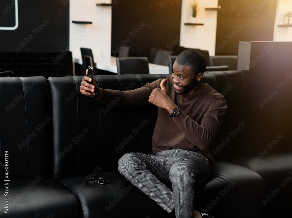 happy young cute fashionably dressed African American man taking a selfie on a smartphone against the background of the dark interior of the cafe