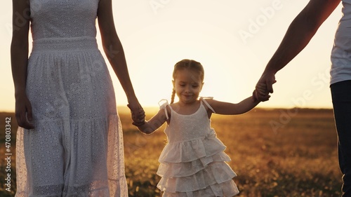 happy baby smiling at sunset with parents, mother, father and child walk in the sun, hold kid by hands, young family, taking care of their little daughter, family walk in evening park in orange light
