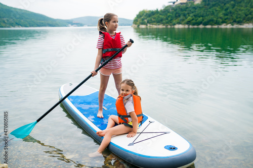 Children in swim life vest swimming on stand up paddle board together. Girls paddleboarding on SUP board on the lake. Active leisure with kids. Family local getaway concept