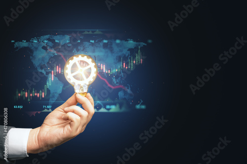 Close up of businessman hand holding lamp with glowing cogwheel with map, forex chart and mock up place on blue background. Conceptual skills, trade and technology concept.