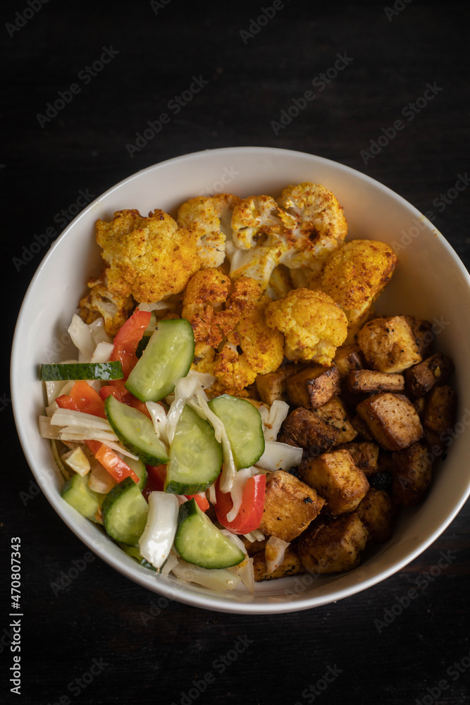 Vegan lunch bowl: baked cauliflower, tofu, tomato and cucumber salad. Dark wooden background. Vegan lunch bowl: baked cauliflower, tofu, tomato and cucumber salad. Dark wooden background.