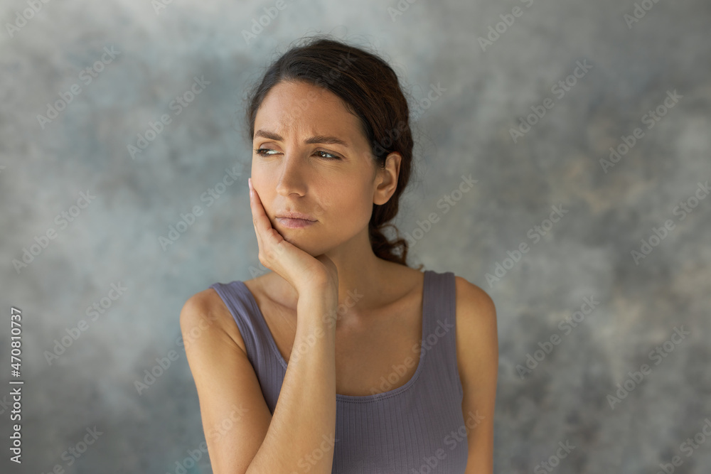 Horizontal studio shot of sad woman suffering from toothache, holding cheek, looking aside with miserable face expression, wearing lilac crop top, isolated over grey textured wall. Human emotions Horizontal studio shot of sad woman suffering from toothache, holding cheek, looking aside with miserable face expression, wearing lilac crop top, isolated over grey textured wall. Human emotions
