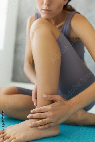 Crop shot of pretty young woman traumatized her ankle during intensive workout at gym, sitting on floor, holding and stroking her injured leg, feeling pain. Sport and its implications