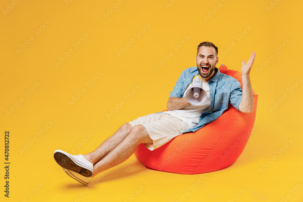 Full size young happy promoter man 20s wearing blue shirt sit in bag chair hold scream in megaphone announces discounts sale Hurry up spread hand isolated on plain yellow background studio portrait
