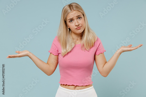 Young uncertain caucasian blonde woman 20s wearing casual pink t-shirt shrugging shoulders looking puzzled, have no idea, nothing to say isolated on plain pastel light blue background studio portrait
