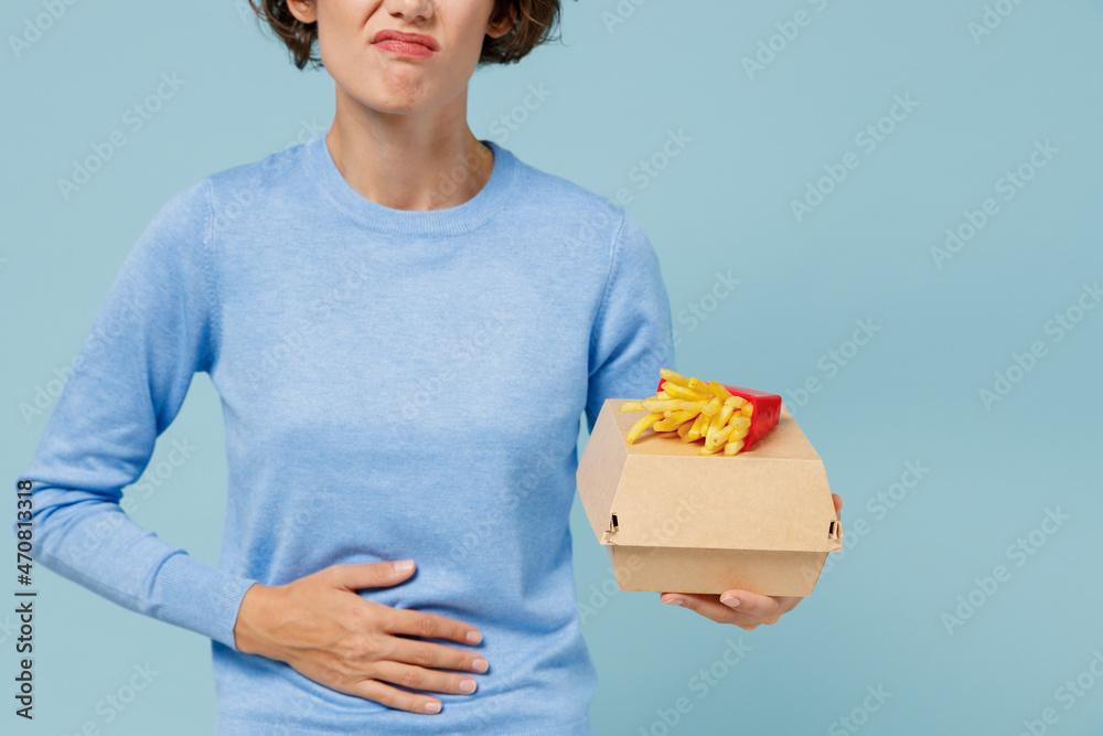 Cropped young hungry sick woman wearing sweater hold a box with fastfood french fries potato burger put hand on belly isolated on plain pastel light blue background People lifestyle junk food concept Cropped young hungry sick woman wearing sweater hold a box with fastfood french fries potato burger put hand on belly isolated on plain pastel light blue background People lifestyle junk food concept