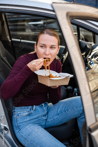 Young blonde girl eating delicious Chinese noodles from the box with chopsticks, sitting in the car. Takeout food. Wok noodle concept