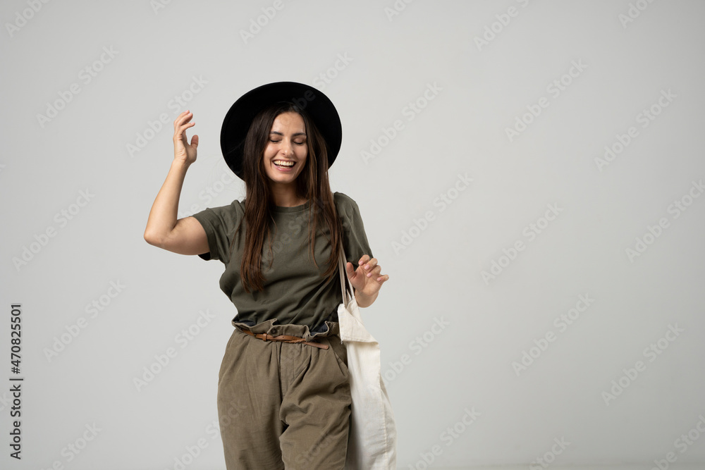 Brunette woman in green t-shirt and black hat holding cotton shopper bag with vegetables, products in white room. Eco friendly shopping bags. Zero waste, plastic free concept.