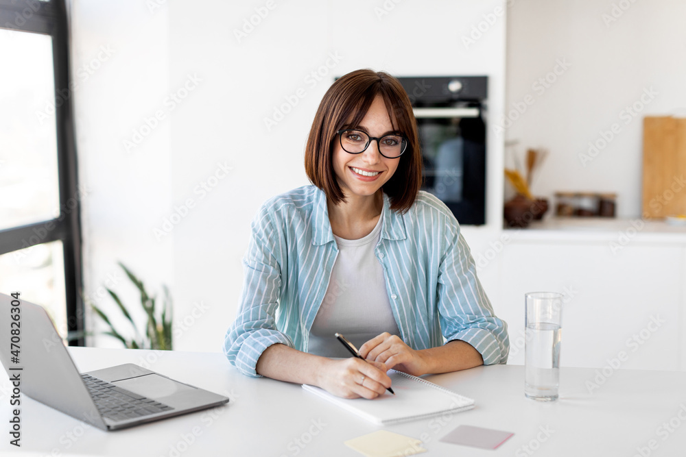 Distance education. Happy woman studying with laptop, taking notes and smiling to camera in kitchen interior Distance education. Happy woman studying with laptop, taking notes and smiling to camera in kitchen interior