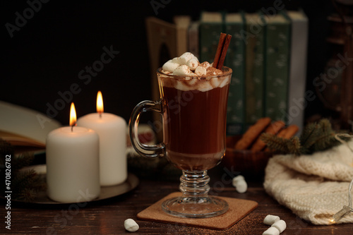 Cocoa or hot chocolate with marshmallows and a cinnamon stick in a tall glass glass. Against the background of books, burning candles and a knitted plaid. Close-up