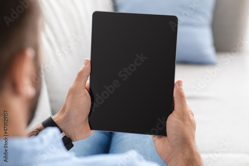 Middle-aged man sits on couch, resting and looking at tablet with blank screen, watch video or ad