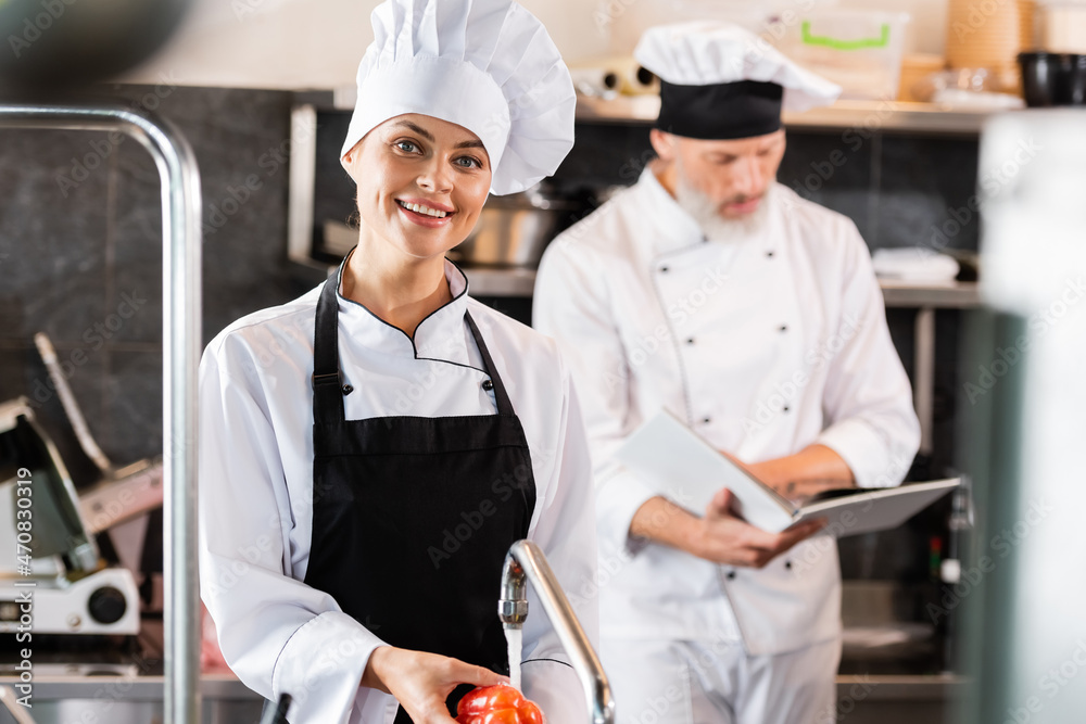 Smiling chef looking at camera while washing bell pepper near colleague with cookbook in kitchen