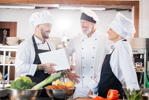 Asian chef holding cookbook and talking to smiling colleagues near food in kitchen