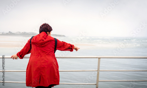 Rear view of a reflective woman red trench coat contemplating the sea on a beach on a summer vacation, outdoors. Middle-aged travel lifestyle. photo