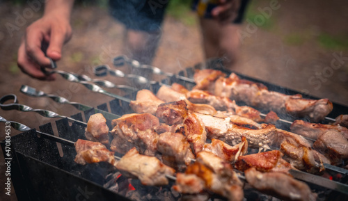 Man hand straightens the skewer with barbecue delicious grilled meat on brazier outside at summertime. Person preparing pork steaks with smoke during picnic at the nature. Closeup view at hot BBQ