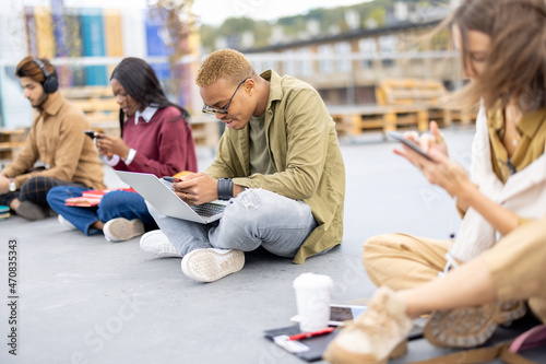 Row of multiracial students sitting and using smartphones on asphalt at university campus. Concept of education. Remote and e-learning. Idea of student lifestyle. Smiling and focused girls and guys