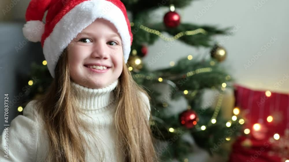 Portrait of Happy little girl in Santa's hat smiling at the New Year on the background of Christmas tree with toy bear. Child feeling excited of getting wished presents, New Year celebration