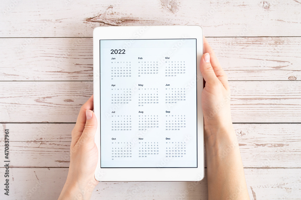 Obraz premium tablet computer with an open app of calendar for 2022 year in a womans hands on a wooden boards background. concept business or to do list goals with technology using. top view, flat lay