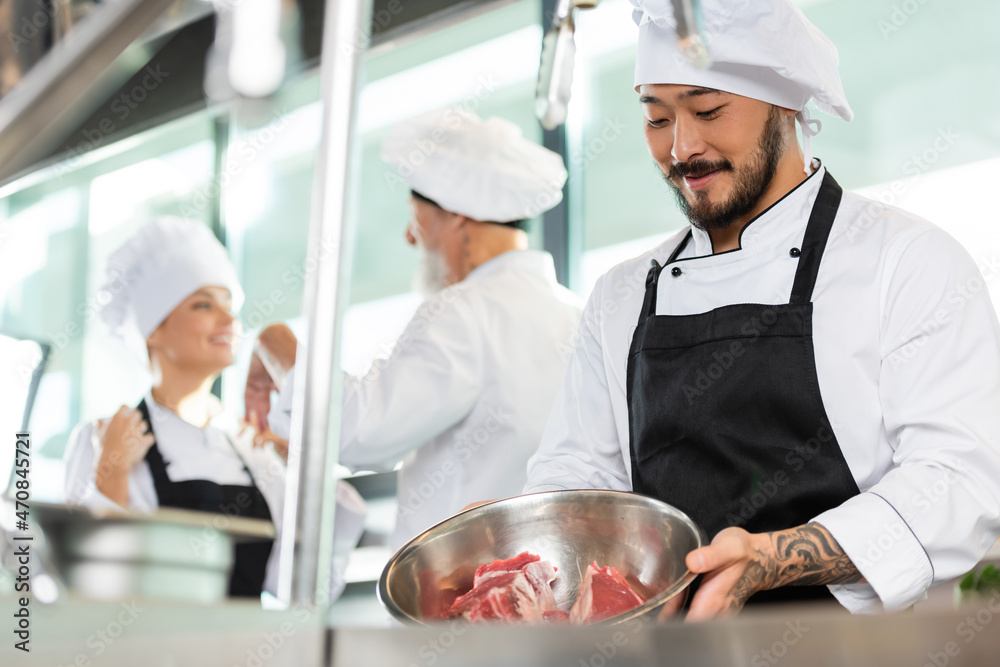 Smiling asian chef holding bowl with meat near colleagues in kitchen Smiling asian chef holding bowl with meat near colleagues in kitchen
