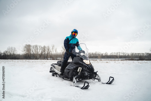 Snowmobile driver stands on his black snowmobile in a snowy area, cross helmet
